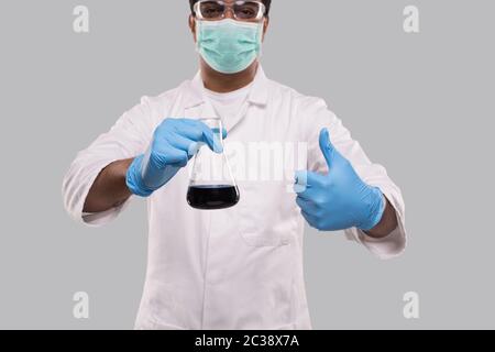 Männlich Arzt Holding Flasche mit Colorfull Flüssigkeit zeigt Daumen nach oben Tragen von medizinischen Maske, Handschuhe und Brille isoliert. Wissenschaft, Medizin, Virenkonzept Stockfoto