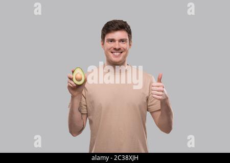 Mann zeigt Avocado und Daumen nach oben isoliert. Avocado in zwei Hälften schneiden Stockfoto