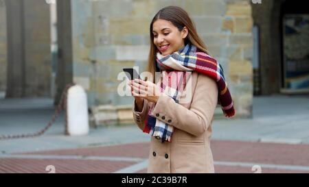 Glücklich lächelnde Frau mit Mantel und Schal mit Smart Phone im Freien in der Stadt. Stockfoto