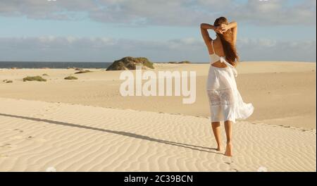 Panoramablick auf die Fahne Ansicht der schönen Frau mit weißem Kleid Wandern auf Dünen der Wüste bei Sonnenuntergang. Mädchen gehen auf goldenen Sand auf Corralejo Dunas, Fuerteven Stockfoto