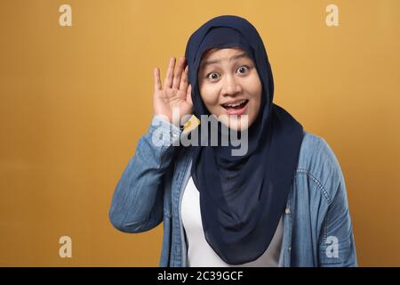 Asiatische muslimische Frau trägt Hijab lächelnd während dabei, Geste zu hören, schockiert überrascht Ausdruck Klatsch zu hören, vor gelbem Hintergrund Stockfoto