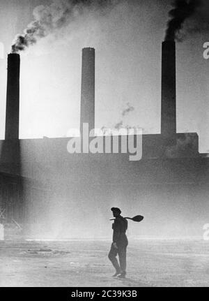 Ein Arbeiter geht auf dem Industriegelände. Im Hintergrund die Schornsteine der Fabrik. Stockfoto