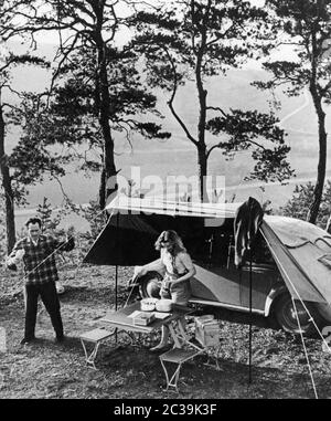 Ein Paar, das während eines Campingurlaugs Camp aufstellend. Eine Plane, die sie über ihr Auto gespannt haben, dient als Baldachin und Schatten. Stockfoto