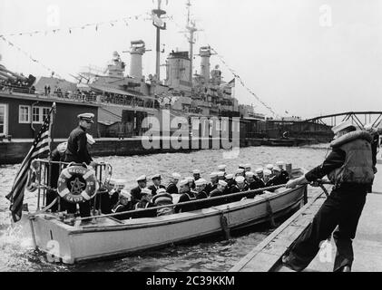 Ein Beiboot des schweren Kreuzfahrtschiffes USS Macon (CA-132, im Hintergrund zu sehen), legt gerade am Steg fest. Matrosen und Offiziere der Besatzung sitzen im Boot. Anlass ist ein Besuch der US Navy in Hamburg in den 1950er Jahren. Stockfoto