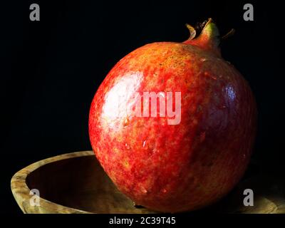 Ein einzelner gewaschene Granatapfel mit Wassertröpfchen in einer Holzschale vor schwarzem Hintergrund Stockfoto