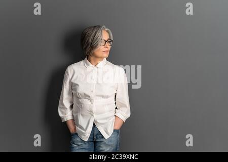 Schöne reife Dame in Brille steht Hände in Taschen Blick auf Seite. Elegante grauhaarige Frau in weißem Hemd auf grauem Hintergrund. Speicherplatz kopieren für Stockfoto