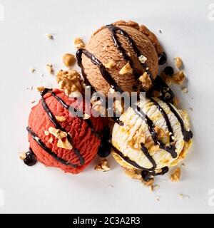 Drei verschiedene aromatisiert Schaufeln des gefrorenen italienisches Eis gekrönt mit beträufelt Schokolade und Nüssen von oben gesehen auf Weiß Stockfoto