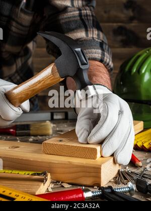 Close-up. Tischler mit seinen Händen durch Handschuhe geschützt, mit Hammer und Nägel behebt ein Holzbrett. Bauwirtschaft, die es selbst tun. Holz- arbeiten Stockfoto