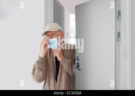 Mann mit Kappe, der seine schützende medizinische Maske anlegt, als er das Haus verlässt. Er geht eine halb geöffnete Tür in einem Gebäude hinaus. Coronavirus Covid-19 Pandemie machen Stockfoto