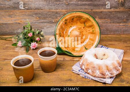 Ein Gugelhupf mit seiner Keramik-Backform und zwei Tassen Kaffee und ein Feld Blumen Geburtstagsstrauß auf einem Holztisch mit einem hölzernen Hintergrund. Und ein Stockfoto