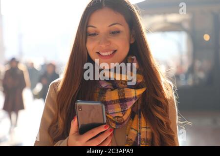 Nahpositive junge Frau erhält gute Nachrichten auf ihrem Telefon, während sie auf der Straße mit verschwommenen Menschenmassen auf dem Hintergrund spazieren geht Stockfoto