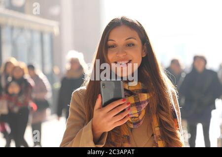 Positiv überraschte junge Frau erhält gute Nachrichten auf ihrem Telefon, während sie auf der Straße mit verschwommenen Menschenmassen auf dem Hintergrund spazieren geht Stockfoto