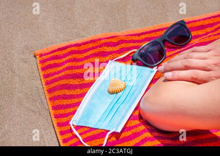 Nahaufnahme eines kaukasischen Mannes, der mit dem Gesicht nach unten auf einem bunten orangefarbenen Handtuch am Strand liegt, neben seiner Sonnenbrille, einer Muschel und einer blauen OP-Maske Stockfoto