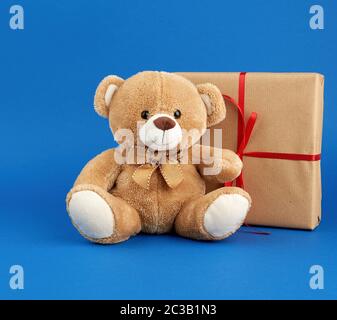 Beigefarbener Teddybär und eine Schachtel in braunem Kraftpapier verpackt und mit rotem Band, festlicher Kulisse für Valentinstag und Geburtstag verbunden Stockfoto