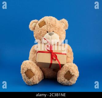 Der große beige Teddybär hält eine Schachtel in braunem Papier und mit rotem Band, festlicher Kulisse für Valentinstag und Geburtstag Stockfoto