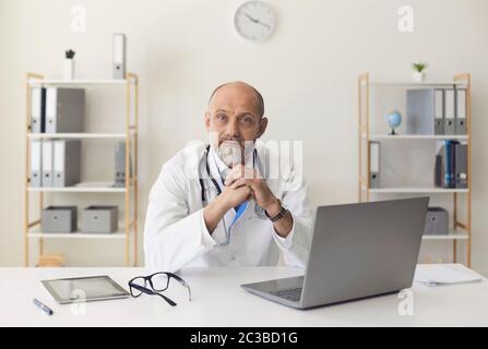 Seriöse Senior Arzt Blick auf die Kamera, während an einem mit Laptop in einer medizinischen Klinik Büro sitzen. Stockfoto