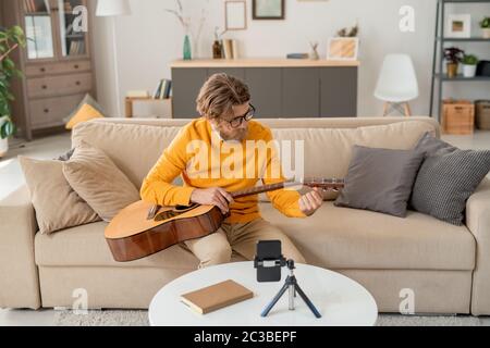 Zeitgenössischer junger Mann in Jeans und gelber Pullover sitzt auf der Couch und zieht Gitarrensaiten vor der Smartphone-Kamera zu Hause Stockfoto