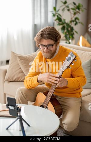 Happy bärtig blonde Kerl mit Gitarre in Smartphone-Kamera suchen, während auf der Couch sitzen und lehren Online-Publikum, wie man Musik zu machen Stockfoto