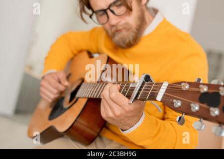 Bärtiger junger Musiker mit Saiteninstrument, das Musik macht oder akustische Gitarre in häuslicher Umgebung stimmt, bevor er es spielt Stockfoto