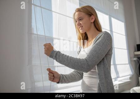 Hübsche, junge Frau, die Senkung der Innenbereich Jalousien/Rollos in ihrer modernen Interieur Wohnung Stockfoto