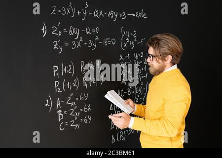 Bärtiger Lehrer in Casualwear Umschreiben Formeln auf Tafel aus seinem Copybook, bevor er Aufgabe zu seinem Online-Publikum Stockfoto