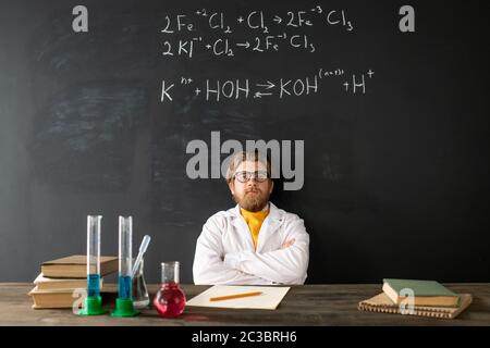 Junger zeitgenössischer Chemielehrer in Whitecoat, der während des Online-Unterrichts gegen Tafel mit chemischen Formeln seine Arme auf der Brust kreuzt Stockfoto