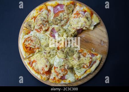 Pizza mit Mozzarella, Salami, Tomaten, Bananen, ohne ein einziges Stück, steht auf einem Holzbrett auf einem schwarzen Hintergrund. top Aussicht Stockfoto