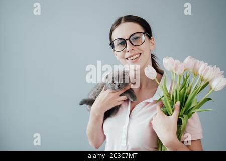 Studio Portrait von jungen Erwachsenen hübsches Mädchen mit Brille in Rosa T-Shirt gekleidet hält Bouquet von frischen Tulpen und ihre kleine Katze Stockfoto