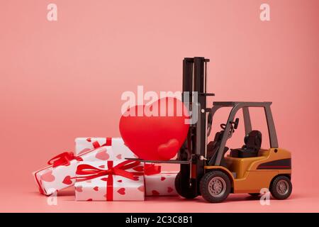 Gabelstapler, rotes Herz und Geschenkboxen auf rosa Hintergrund. Valentinstag. Hochzeitstag Stockfoto