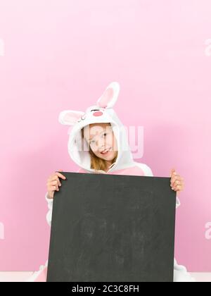 Hübsches blondes Mädchen mit gemütlichem Kaninchen-Kostüm und Tafel posiert im Studio und ist glücklich Stockfoto