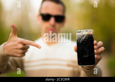 Kerl hält schwarzes Smartphone mit defekten Display. Kaputter Bildschirm des modernen rahmenlosen Telefon. Kerl weinend mit geschlossenen Augen Stockfoto