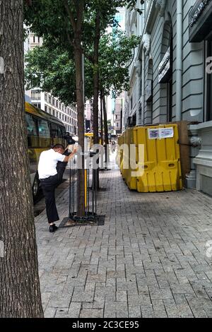 Ein Mann, der sein Bein sehr hoch streckt, während er neben dem Baum auf dem Bürgersteig steht, ein weißes Hemd und eine schwarze Hose trägt. Stockfoto