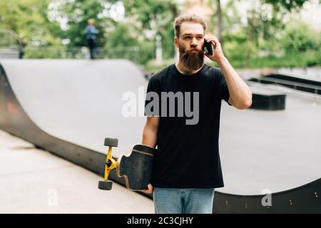 Der junge Mann mit Skateboard spricht an einem sonnigen Tag mit dem Handy in einem Skatepark. Stockfoto