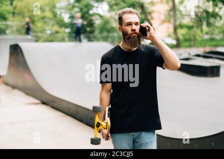 Der junge Mann mit Skateboard spricht an einem sonnigen Tag mit dem Handy in einem Skatepark. Stockfoto
