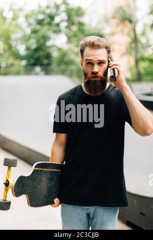 Der junge Mann mit Skateboard spricht an einem sonnigen Tag mit dem Handy in einem Skatepark. Stockfoto