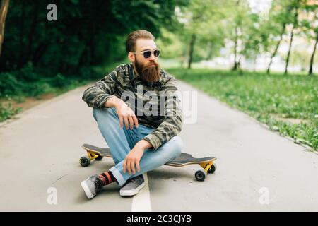 Junge bärtige Mann Longboarder in lässiger Kleidung sitzt auf dem Longboard oder Skateboard im Freien. Urban, Subkultur, Skateboarding Konzept Stockfoto