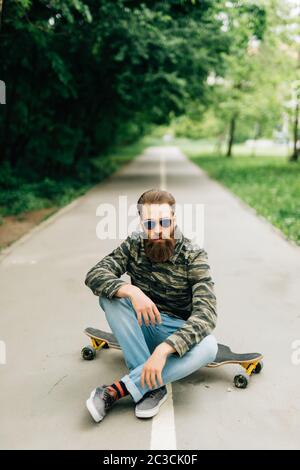 Junge bärtige Mann Longboarder in lässiger Kleidung sitzt auf dem Longboard oder Skateboard im Freien. Urban, Subkultur, Skateboarding Konzept Stockfoto