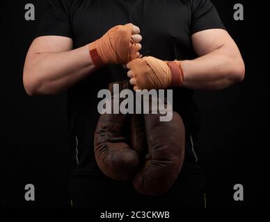 Muskelsportler in einer schwarzen Uniform hält sehr alte braune Boxhandschuhe in der Hand, seine Hände sind mit einem orangefarbenen elastischen Sportverband verbunden, Blac Stockfoto