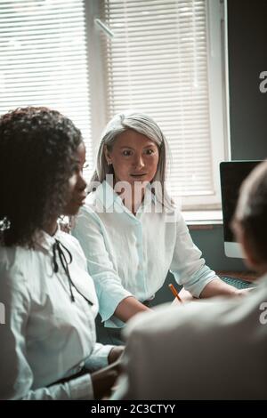 Überraschte Geschäftsfrau macht große Augen, während sie mit Kollegen während der Diskussion kommuniziert. Konzentrieren Sie sich auf grauhaarige asiatische Frau am Schreibtisch sitzen Stockfoto