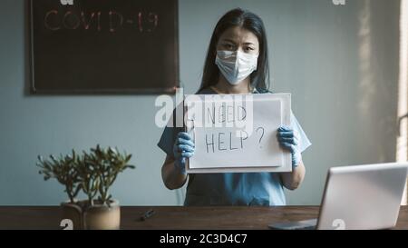 Seriöse Arzt bietet Hilfe an. Asiatische Frau in Schutzuniform mit langen Haaren schaut auf Kamera stehen in ihrem medizinischen Büro. Getöntes Bild Stockfoto