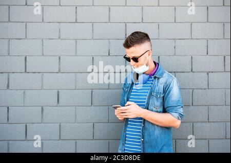 Junger Mann mit Kopfhörern um den Hals und tiefer OP-Maske nach Coronavirus Ausbruch - trendy Kerl beobachten Video auf Smartphone oder Chat - Stockfoto