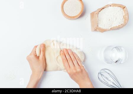 Draufsicht Frauenhände Kneten von Teig neben Mehl und Wasser Einfache Zutaten für Brot, Pizza oder Pasta auf weißem Tisch. Stockfoto