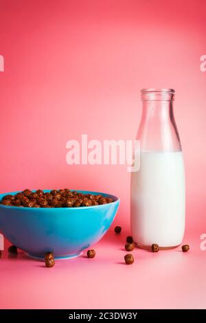 Milch in einer Glasflasche und Frühstücksflocken aus Schokolade in einer blauen Schüssel auf rosa Hintergrund. Selektiver Fokus Stockfoto