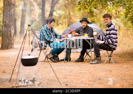 Gruppe von Leuten, die am Tisch sitzen und Essen auf Feuer gekocht essen. Lagergut. Gruppe von jungen Menschen in Freizeitkleidung Ruhe im Herbst Wald Stockfoto