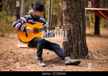 Junge attraktive Mann im Pullover spielen Gitarre im Wald sitzen auf Schaukel mit Hängematte auf Hintergrund. Lässiger Mann, Lifestyle. Stockfoto