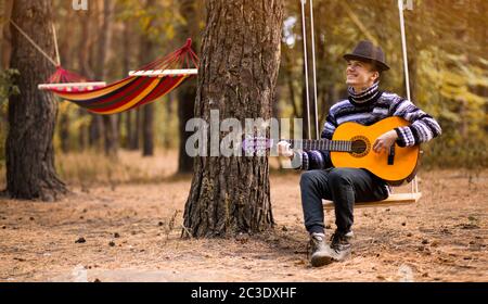 Junge attraktive Mann im Pullover spielen Gitarre im Wald sitzen auf Schaukel mit Hängematte auf Hintergrund. Lässiger Mann, Lifestyle. Stockfoto