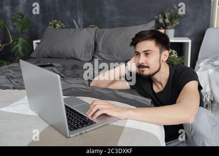 Inhalt junger Mann mit Schnurrbart, der Laptop im Bett benutzt, während er über eine Videokonferenz-App chattet, während er sich selbst isoliert Stockfoto