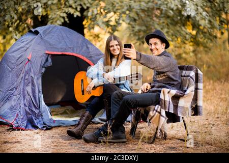 Glückliches junges Paar macht Selfie mit Smartphone und Spaß Camping spielen Gitarre im Herbst Wald in der Nähe Zelt Stockfoto