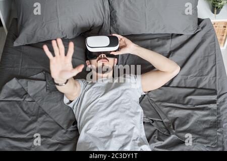 Oben Ansicht des jungen bärtigen Mann eingetaucht in virtuelle Realität liegend auf dem Bett und Video in VR-Brille Stockfoto