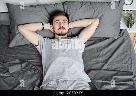 Oben Ansicht von nachdenklichen jungen bärtigen Mann, der Hände hinter dem Kopf hält, während er sich im Bett während der Quarantäne entspannt Stockfoto